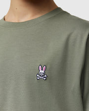 Close-up of a person wearing the KIDS CLASSIC CREW NECK TEE - B0U014B200 by Psycho Bunny in olive green. Crafted from 100% Pima cotton, the shirt features a small embroidered logo on the chest, showcasing a skull and crossbones with bunny ears in pink. Perfect for back-to-school outfits, it’s one of those kids' classic crew neck tee shirts they'll love.