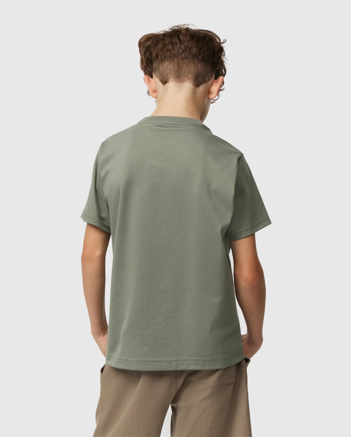A young boy with short, curly hair is standing with his back facing the camera. He is wearing an olive green Psycho Bunny KIDS CLASSIC CREW NECK TEE - B0U014B200 made from 100% Pima cotton and light brown pants, perfect for back-to-school outfits. The background is plain white.