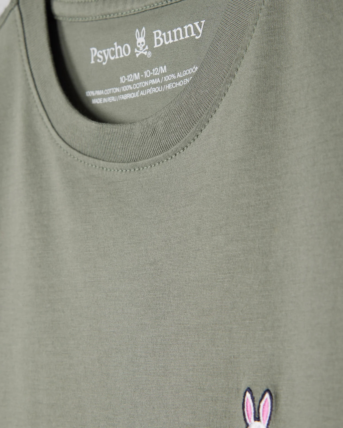 Close-up of a khaki green Kids Classic Crew Neck Tee (B0U014B200) by Psycho Bunny, highlighting the neckline and chest area. Crafted from 100% Pima cotton, it features the brand's iconic bunny skull and crossbones logo embroidered in pink and white on the left side, with size and material information printed inside the collar. An ideal choice for back-to-school outfits.