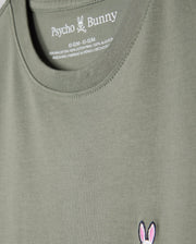 Close-up of a khaki green Kids Classic Crew Neck Tee (B0U014B200) by Psycho Bunny, highlighting the neckline and chest area. Crafted from 100% Pima cotton, it features the brand's iconic bunny skull and crossbones logo embroidered in pink and white on the left side, with size and material information printed inside the collar. An ideal choice for back-to-school outfits.