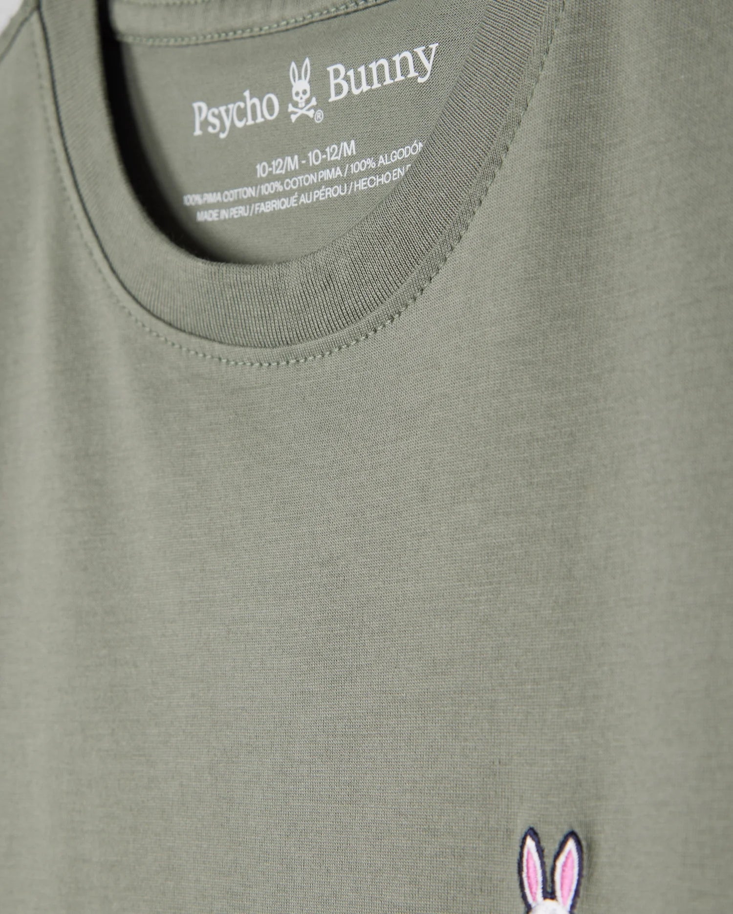 Close-up of a khaki green Kids Classic Crew Neck Tee (B0U014B200) by Psycho Bunny, highlighting the neckline and chest area. Crafted from 100% Pima cotton, it features the brand's iconic bunny skull and crossbones logo embroidered in pink and white on the left side, with size and material information printed inside the collar. An ideal choice for back-to-school outfits.