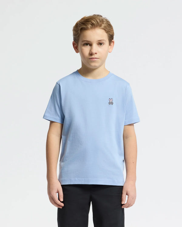 A young boy with short hair stands against a plain background wearing the Psycho Bunny KIDS CLASSIC CREW NECK TEE in light blue, crafted from soft Pima cotton with an embroidered Bunny logo on the chest, paired with sleek black pants.