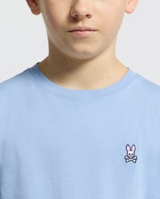 A child wears a Psycho Bunny light blue KIDS CLASSIC CREW NECK TEE (B0U014B200) made of Pima cotton, featuring an embroidered bunny logo with crossed bones. The focus is on the neck and upper chest area.