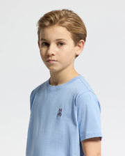 A young boy with light brown hair stands against a plain white background, wearing a Psycho Bunny Kids Classic Crew Neck Tee in light blue Pima cotton with an embroidered bunny logo.