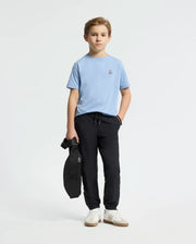 A young boy stands against a plain background, wearing the light blue Psycho Bunny Kids Classic Crew Neck Tee (B0U014B200) with black pants. He holds a black bag and sports white sneakers as an embroidered Bunny logo subtly adorns his tee.
