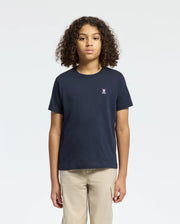 A person with curly hair wears a Psycho Bunny KIDS CLASSIC CREW NECK TEE in navy blue, made from 100% Pima cotton with a small white logo on the chest. They're styled with beige pants against a plain white background—a timeless back-to-school look.