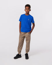 A young boy stands confidently with his hands in his pants pockets, wearing a bright blue KIDS CLASSIC CREW NECK TEE by Psycho Bunny, crafted from soft Pima cotton. Beige jogger pants and black sneakers complete his look against a plain white background.
