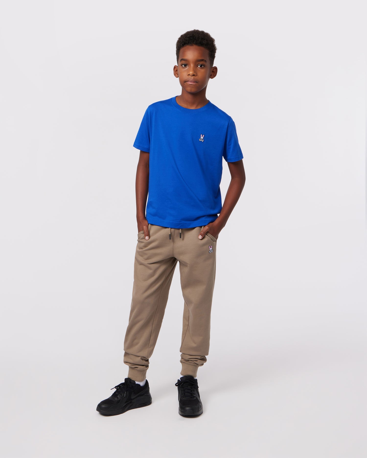 A young boy stands confidently with his hands in his pants pockets, wearing a bright blue KIDS CLASSIC CREW NECK TEE by Psycho Bunny, crafted from soft Pima cotton. Beige jogger pants and black sneakers complete his look against a plain white background.