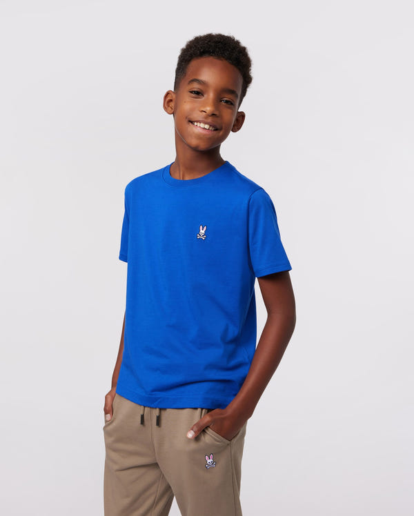 A smiling boy in a Psycho Bunny KIDS CLASSIC CREW NECK TEE (B0U014CRPC), featuring a small rabbit logo, stands casually with one hand in his pocket. He stylishly pairs it with beige pants that have a matching logo, set against a plain white background.