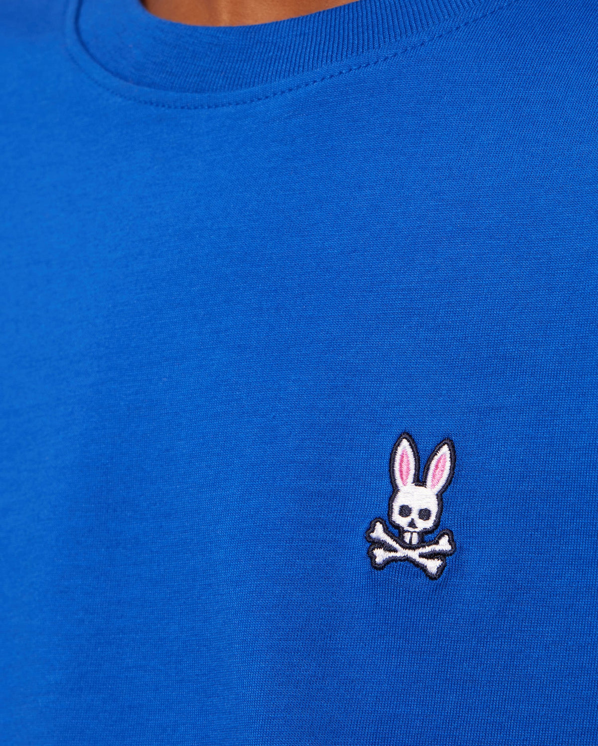 A close-up of an individual wearing the Psycho Bunny KIDS CLASSIC CREW NECK TEE in blue Pima cotton, adorned with a small embroidered design of a white rabbit skull with pink ears above crossed bones on the chest.