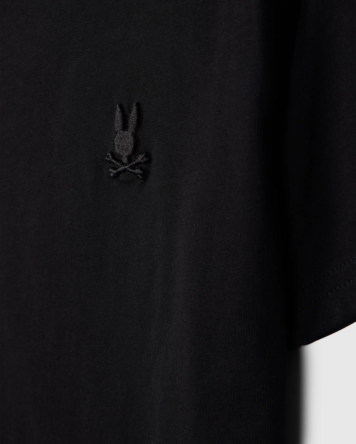 Close-up of a Psycho Bunny Kids Embroidered Classic Tee, product code B0U020AR00, crafted from luxurious Peruvian Pima cotton. It features a small bunny head with crossed bones design on the left chest and is displayed against a plain background for a timeless silhouette.