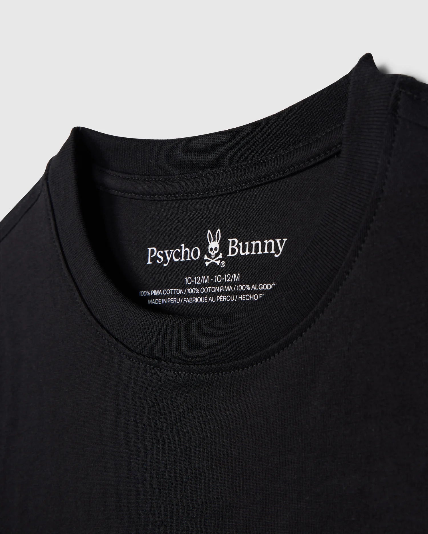 Close-up image of a black T-shirt neckline with a label showing the brand name "Psycho Bunny" alongside its bunny logo, and the size 10-12 M. The KIDS EMBROIDERED CLASSIC TEE - B0U020AR00 is made of 100% Peruvian Pima cotton, ensuring ultimate comfort and quality. This piece showcases a timeless silhouette and is proudly made in Peru.