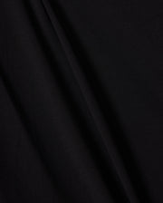A close-up image of the smooth, black Peruvian Pima cotton fabric used in the KIDS EMBROIDERED CLASSIC TEE - B0U020AR00 by Psycho Bunny. The texture appears soft and slightly matte, with subtle folds and creases creating faint shadows that emphasize its draping and natural curvature, evoking a timeless silhouette.