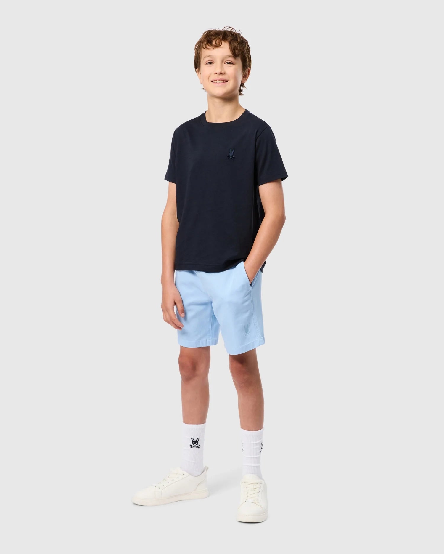 A smiling boy stands against a plain background, wearing a dark navy KIDS EMBROIDERED CLASSIC TEE - B0U020AR00 by Psycho Bunny made from Peruvian Pima cotton and light blue shorts. He has white socks with a black emblem and is wearing white sneakers. His right hand is in his pocket, and an embroidered chest Bunny adds a charming touch to his outfit.