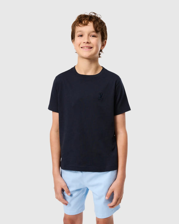A smiling young boy with short, curly brown hair is wearing a navy blue KIDS EMBROIDERED CLASSIC TEE - B0U020AR00 from Psycho Bunny, made from Peruvian Pima cotton and featuring an embroidered chest Bunny on the left side. He pairs it with light blue shorts and stands against a plain, light gray background.