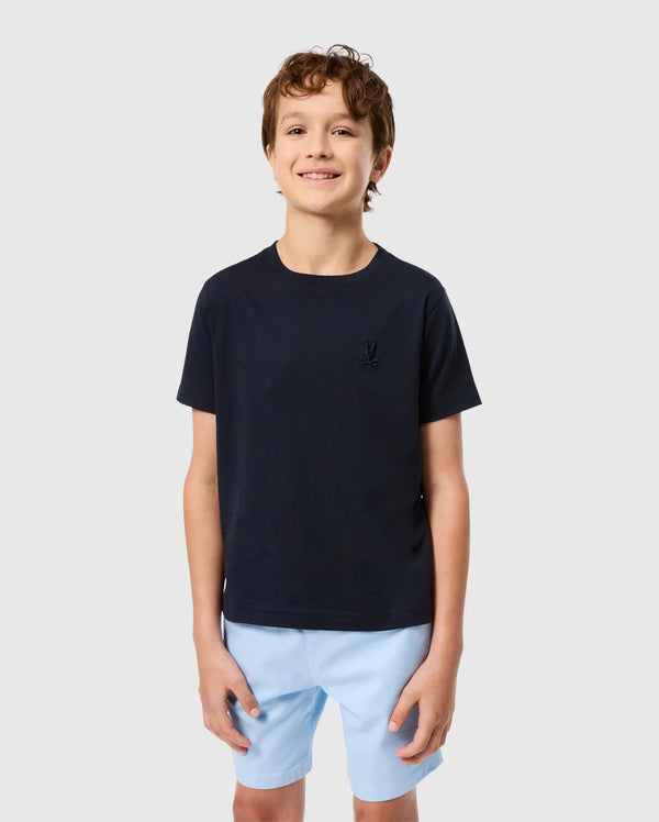 A smiling young boy with short, curly brown hair is wearing a navy blue KIDS EMBROIDERED CLASSIC TEE - B0U020AR00 from Psycho Bunny, made from Peruvian Pima cotton and featuring an embroidered chest Bunny on the left side. He pairs it with light blue shorts and stands against a plain, light gray background.