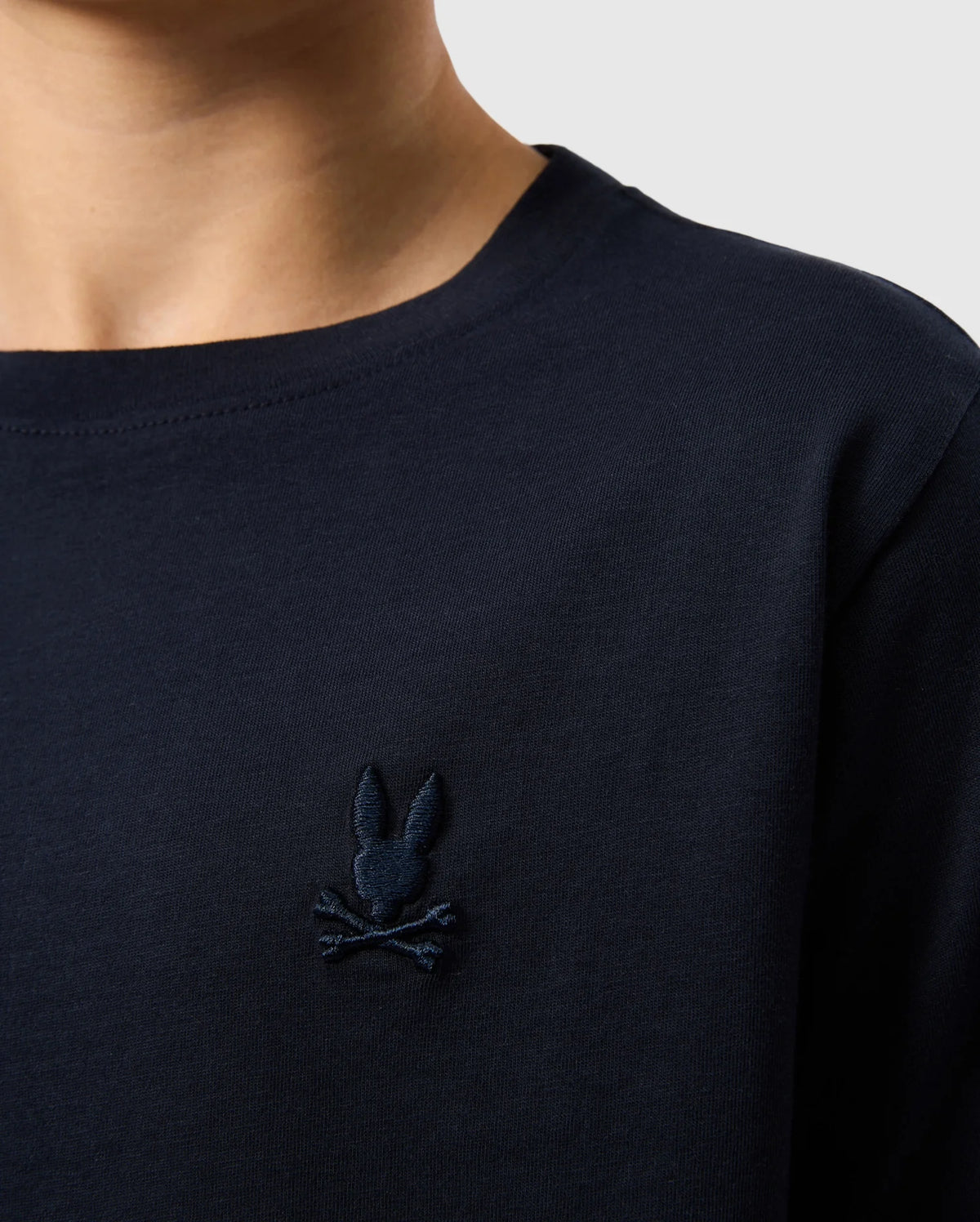 Close-up of a person wearing the Kids Embroidered Classic Tee - B0U020AR00 by Psycho Bunny, made from soft Peruvian Pima cotton. The shirt is a dark navy blue and features an embroidered chest Bunny logo, consisting of a bunny head with long ears above two crossed bones, all in the same navy blue color as the shirt. The background is a plain light gray.