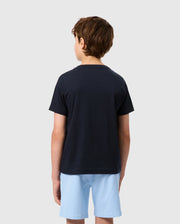 A boy with short brown hair, dressed in a Psycho Bunny KIDS EMBROIDERED CLASSIC TEE - B0U020AR00 and light blue shorts, is standing and facing away from the camera against a plain white background.