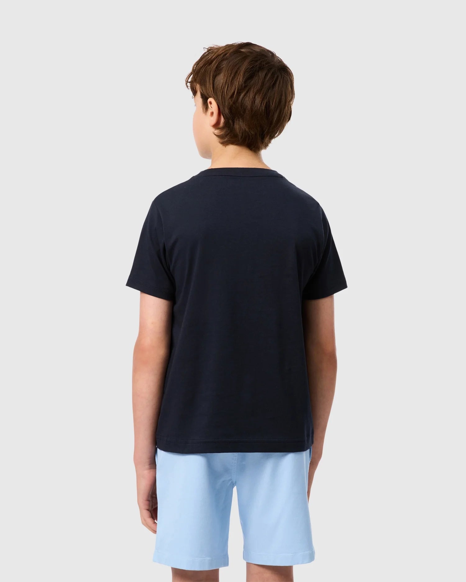 A boy with short brown hair, dressed in a Psycho Bunny KIDS EMBROIDERED CLASSIC TEE - B0U020AR00 and light blue shorts, is standing and facing away from the camera against a plain white background.