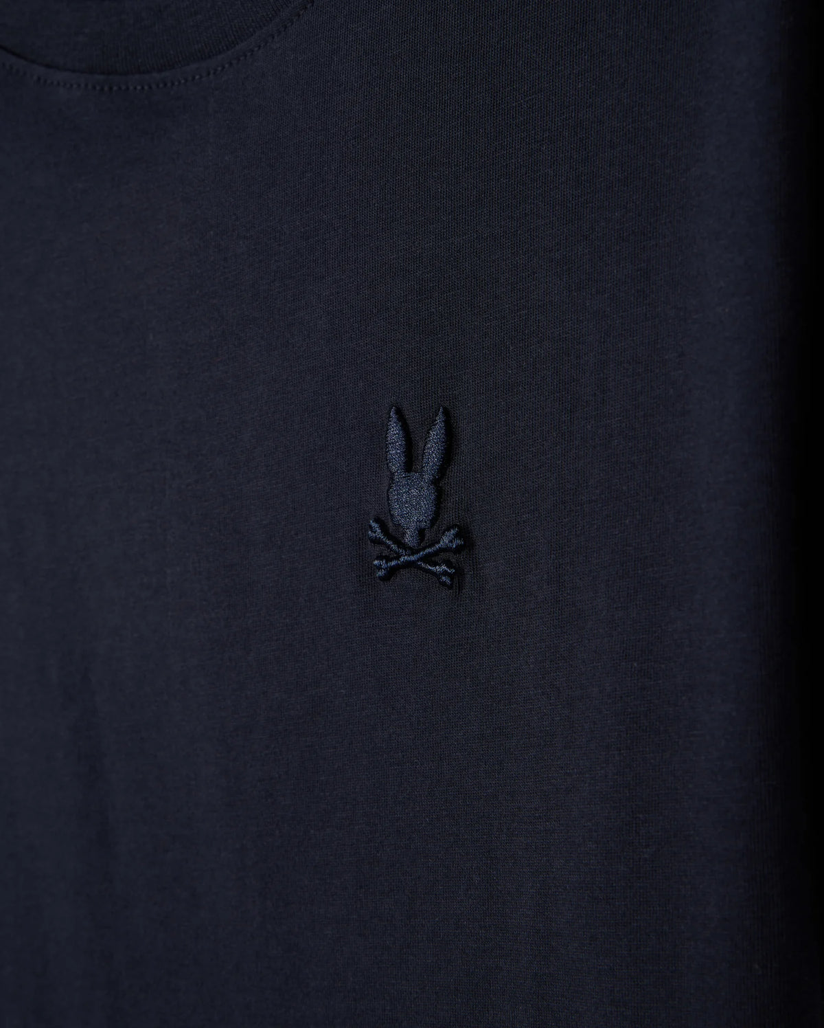 A close-up of the Kids Embroidered Classic Tee by Psycho Bunny, showcasing a dark-colored fabric with a black embroidered design featuring a bunny head with long ears above crossed bones, akin to a skull and crossbones, crafted from luxurious Peruvian Pima cotton.