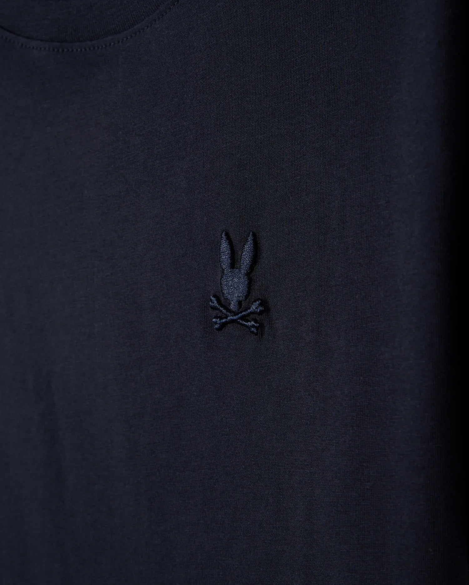 A close-up of the Kids Embroidered Classic Tee by Psycho Bunny, showcasing a dark-colored fabric with a black embroidered design featuring a bunny head with long ears above crossed bones, akin to a skull and crossbones, crafted from luxurious Peruvian Pima cotton.