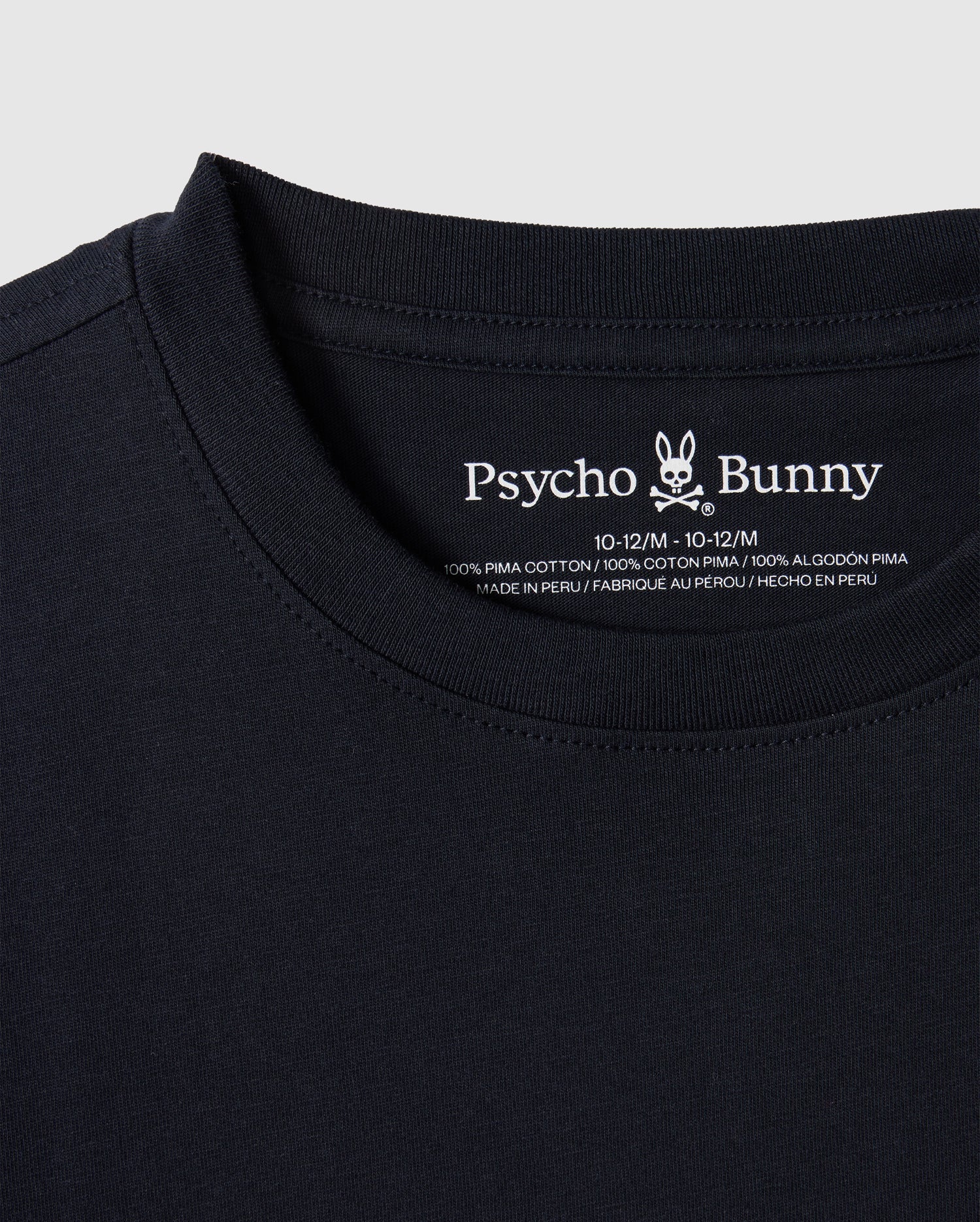 Close-up of the KIDS EMBROIDERED CLASSIC TEE - B0U020AR00 in black, featuring the Psycho Bunny brand logo and text on the inside neck area. It boasts an embroidered chest bunny, and the label confirms it's made of 100% Peruvian Pima cotton, manufactured in Peru.