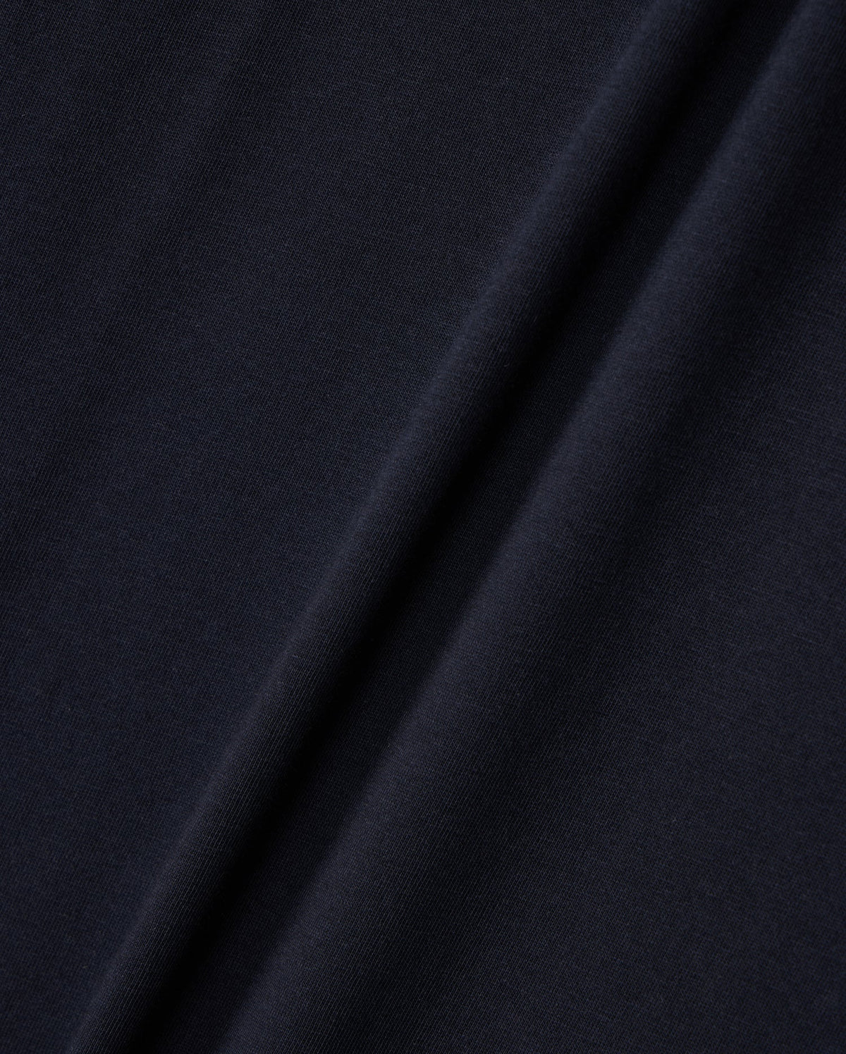 Close-up image of a dark navy blue fabric with a diagonal fold running from the bottom left to the top right. The texture, reminiscent of Peruvian Pima cotton, appears smooth and slightly matte, with subtle variations in color due to the lighting and fabric draping. The fabric is from Psycho Bunny's KIDS EMBROIDERED CLASSIC TEE - B0U020AR00.