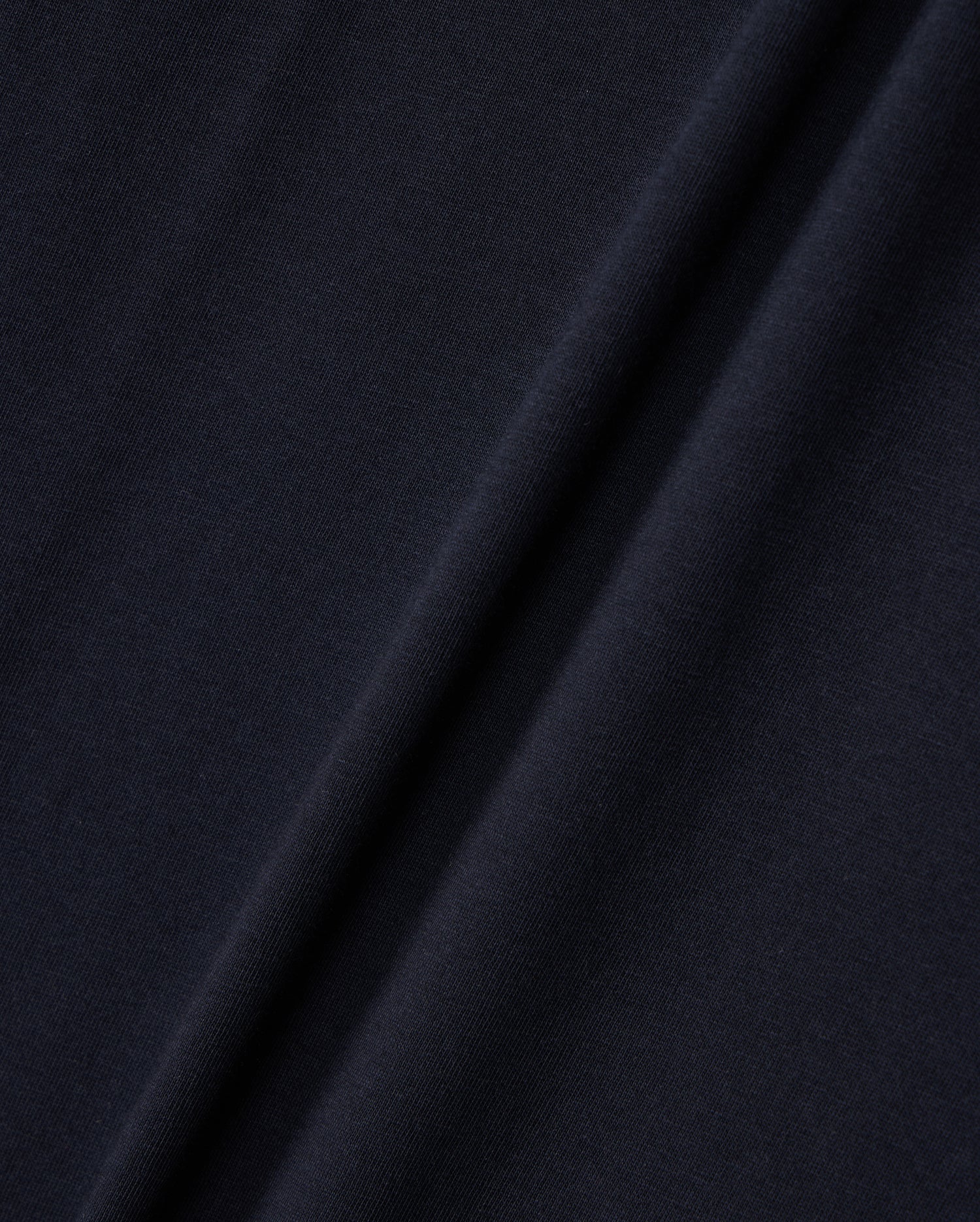 Close-up image of a dark navy blue fabric with a diagonal fold running from the bottom left to the top right. The texture, reminiscent of Peruvian Pima cotton, appears smooth and slightly matte, with subtle variations in color due to the lighting and fabric draping. The fabric is from Psycho Bunny's KIDS EMBROIDERED CLASSIC TEE - B0U020AR00.