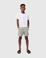 A young boy stands smiling against a plain light grey background. He is wearing the Psycho Bunny KIDS EMBROIDERED CLASSIC TEE - B0U020AR00, made from Peruvian Pima cotton, along with light green shorts and his hands in pockets. His tee features an embroidered chest bunny, adding a playful touch to his relaxed and happy demeanor. Black slides complete his look.