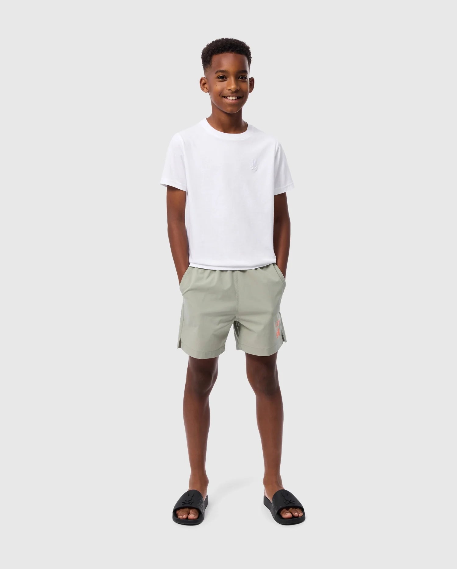 A young boy stands smiling against a plain light grey background. He is wearing the Psycho Bunny KIDS EMBROIDERED CLASSIC TEE - B0U020AR00, made from Peruvian Pima cotton, along with light green shorts and his hands in pockets. His tee features an embroidered chest bunny, adding a playful touch to his relaxed and happy demeanor. Black slides complete his look.