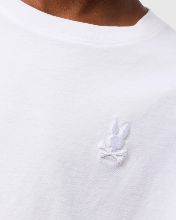 Close-up of a person wearing the white KIDS EMBROIDERED CLASSIC TEE - B0U020AR00 by Psycho Bunny. The visible portion of the Peruvian Pima cotton T-shirt features an embroidered chest bunny head with crossbones below it, stitched in white on the upper chest area. The neckline of the T-shirt is also visible.