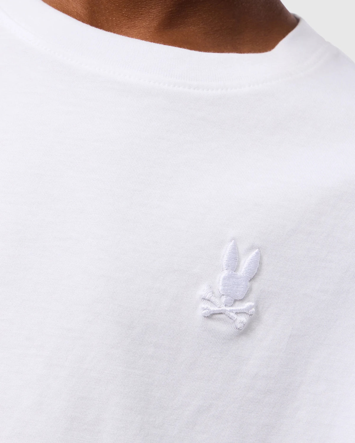 Close-up of a person wearing the white KIDS EMBROIDERED CLASSIC TEE - B0U020AR00 by Psycho Bunny. The visible portion of the Peruvian Pima cotton T-shirt features an embroidered chest bunny head with crossbones below it, stitched in white on the upper chest area. The neckline of the T-shirt is also visible.