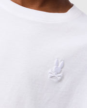 Close-up of a person wearing the white KIDS EMBROIDERED CLASSIC TEE - B0U020AR00 by Psycho Bunny. The visible portion of the Peruvian Pima cotton T-shirt features an embroidered chest bunny head with crossbones below it, stitched in white on the upper chest area. The neckline of the T-shirt is also visible.