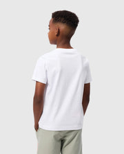 A young boy with short hair is standing with his back towards the camera, wearing a white Psycho Bunny Kids Embroidered Classic Tee - B0U020AR00 made of soft Peruvian Pima cotton and light gray shorts. His hands are tucked into his pockets, and the background is plain and light gray.