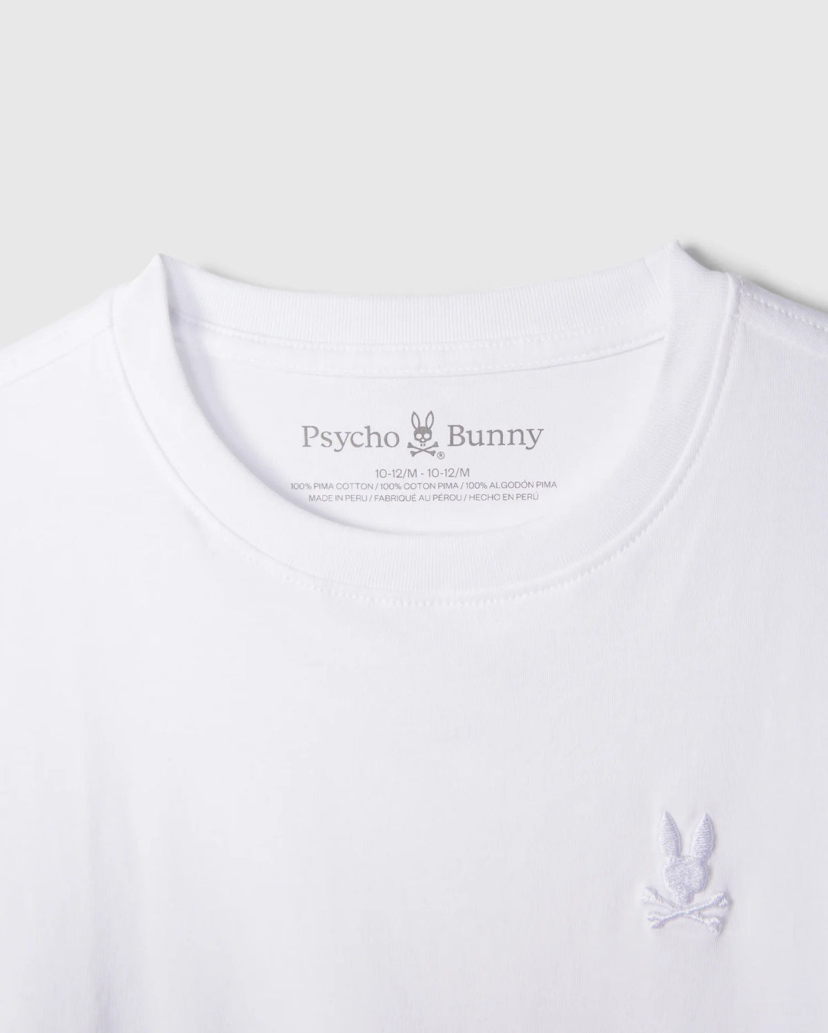 Close-up of a white KIDS EMBROIDERED CLASSIC TEE - B0U020AR00 from Psycho Bunny. This timeless t-shirt showcases a small embroidered Bunny logo on the right side of the chest, featuring a bunny with crossbones underneath. Crafted from luxurious Peruvian Pima cotton, "Psycho Bunny" is printed on the inside neck area along with size and fabric details.