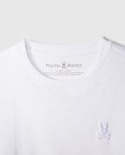 Close-up of a white KIDS EMBROIDERED CLASSIC TEE - B0U020AR00 from Psycho Bunny. This timeless t-shirt showcases a small embroidered Bunny logo on the right side of the chest, featuring a bunny with crossbones underneath. Crafted from luxurious Peruvian Pima cotton, "Psycho Bunny" is printed on the inside neck area along with size and fabric details.