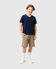 A young person stands smiling in a Psycho Bunny KIDS CLASSIC V NECK TEE - B0U100CRPC, crafted from soft Pima cotton. They pair the navy shirt with beige shorts and white sneakers, while the plain light gray background enhances the casual back-to-school look.