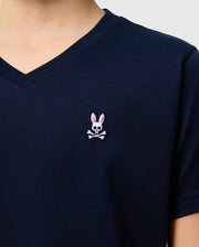 Close-up of a person wearing the Psycho Bunny KIDS CLASSIC V NECK TEE - B0U100CRPC in navy blue, expertly crafted from soft Pima cotton. The shirt features the brand's distinctive skull and crossbones design in white and pink on the chest. The background is a plain light gray, creating an ideal back-to-school vibe.