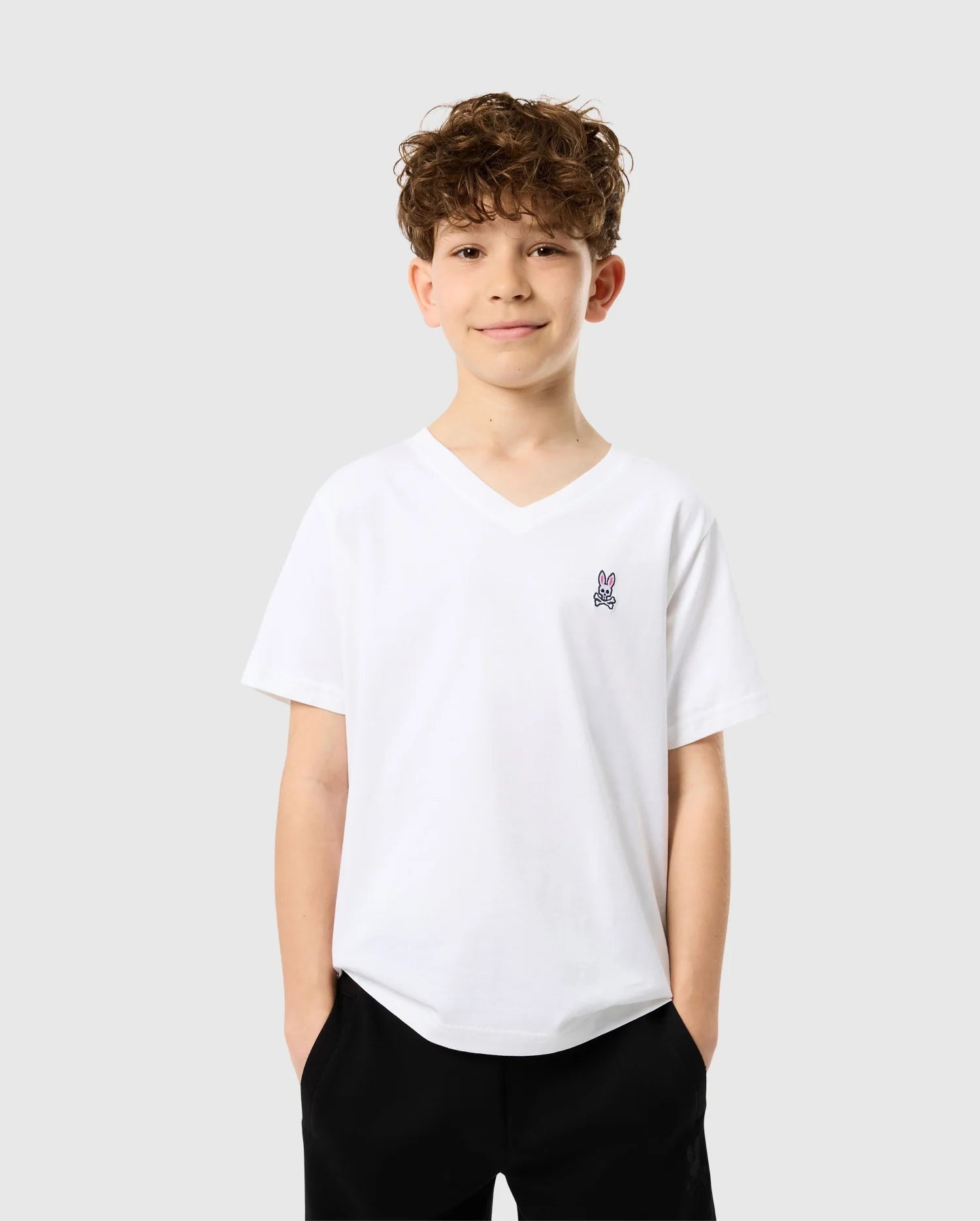 A young boy with curly hair grins as he sports the Psycho Bunny KIDS CLASSIC V NECK TEE - B0U100CRPC. The white V-neck t-shirt, made from soft Pima cotton and featuring a bunny logo on the chest, perfectly complements his back-to-school look. Standing confidently against a plain light gray background, he tucks his hands into the pockets of his dark pants.