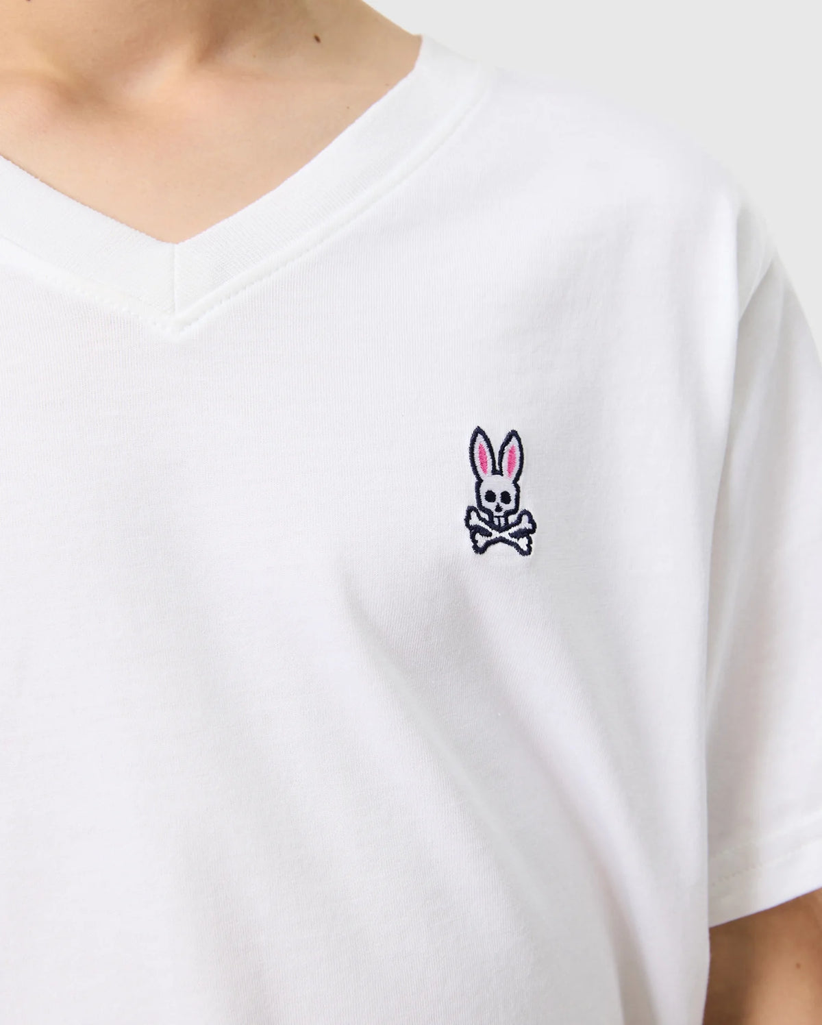 A person is wearing the KIDS CLASSIC V NECK TEE - B0U100CRPC from Psycho Bunny, which is a white Pima cotton shirt featuring a small embroidered logo of a pink-eared bunny with a skull and crossbones design on the chest. The light-colored background complements this back-to-school style.