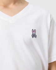 A person is wearing the KIDS CLASSIC V NECK TEE - B0U100CRPC from Psycho Bunny, which is a white Pima cotton shirt featuring a small embroidered logo of a pink-eared bunny with a skull and crossbones design on the chest. The light-colored background complements this back-to-school style.