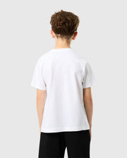 A young boy with curly brown hair stands facing away, wearing a Psycho Bunny KIDS CLASSIC V NECK TEE - B0U100CRPC in white, made of soft Pima cotton, and black shorts. The plain light grey background captures the simplicity and carefree essence of a back-to-school season.