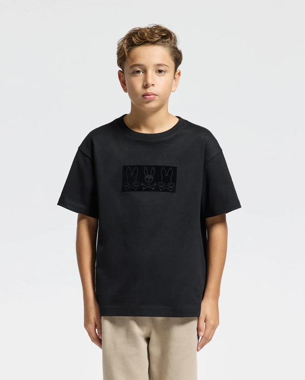 A young boy with short, curly hair stands against a plain background. He is wearing the KIDS ROMEO HEAVY WEIGHT RELAXED FIT TEE - B0U110E200 from Psycho Bunny, crafted from soft pima cotton with a subtle print of three bunnies. His beige pants complement the heavyweight T-shirt, and his expression remains neutral.