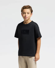 A young boy with curly hair stands against a plain white background wearing a Kids' Romeo Heavy Weight Relaxed Fit Tee by Psycho Bunny, made from soft Pima Cotton. Beige pants complete his look as he rests his hands at his sides, maintaining a neutral expression.
