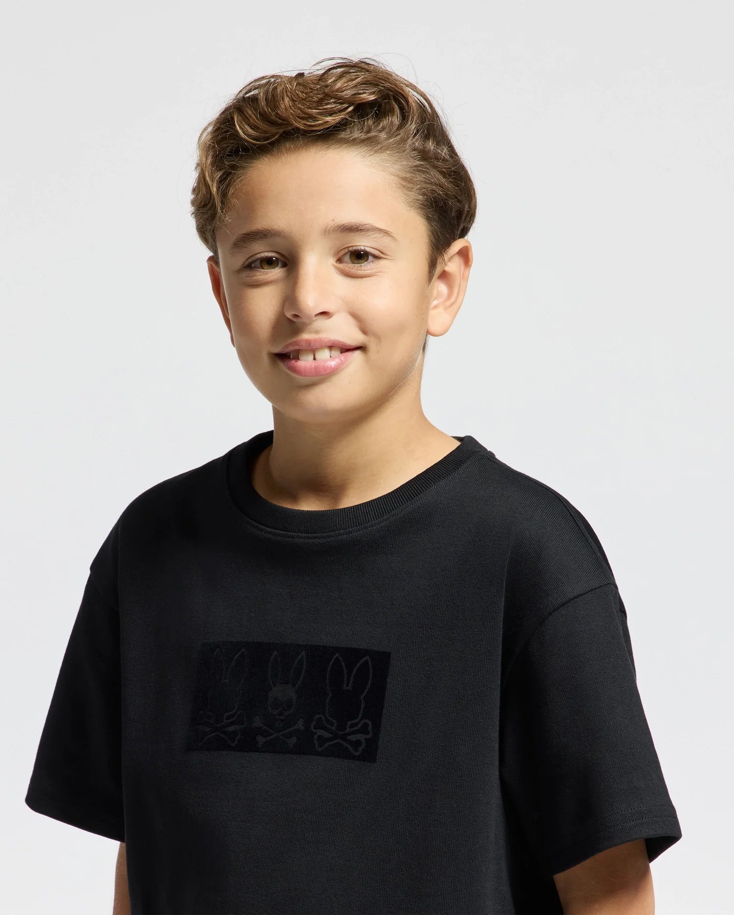 A young boy with short, light brown hair is wearing the Psycho Bunny KIDS ROMEO HEAVY WEIGHT RELAXED FIT TEE - B0U110E200, a black kids' tee adorned with a design of three rabbits on the front. Made from heavyweight Pima cotton, he smiles while standing against a plain, light gray background.