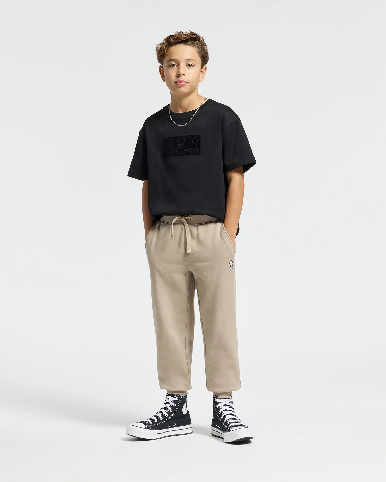 A young boy stands confidently with hands in his pockets, wearing a Psycho Bunny KIDS ROMEO HEAVY WEIGHT RELAXED FIT TEE in black, paired with beige pants and black high-top sneakers. The plain and minimal background highlights his casual yet stylish outfit.