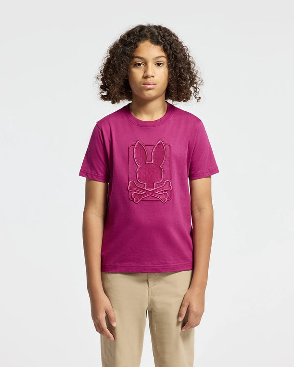 Against a simple background, a young person with curly hair is seen wearing the KIDS PIERCE GRAPHIC TEE - B0U115E200 by Psycho Bunny. This magenta T-shirt, crafted from Kids' Pima Cotton, features an embroidered bunny graphic logo above crossbones and is paired with beige pants.