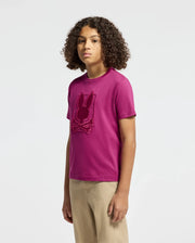 A person with curly hair is wearing the Psycho Bunny Kids Pierce Graphic Tee in magenta, which features an embroidered bunny graphic and crossbones. Against a plain white background, they pair it with beige pants, exuding a playful charm similar to a kids' Pima cotton T-shirt.
