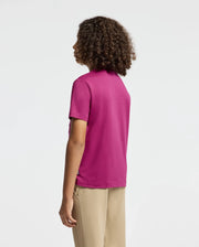 A person with curly hair is standing and facing away, wearing a bright pink KIDS PIERCE GRAPHIC TEE by Psycho Bunny featuring an embroidered bunny graphic, paired with beige pants against a plain, light background.