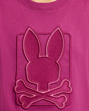 The KIDS PIERCE GRAPHIC TEE - B0U115E200 by Psycho Bunny features a magenta Pima cotton design adorned with a textured emblem. The emblem showcases a stylized bunny graphic above crossed bones, complete with an embroidered design and raised border for a striking three-dimensional effect—perfect for kids who enjoy playful details!